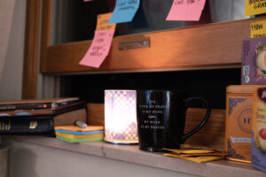 Two mugs sit on a windowsill surrounded by books, tea boxes, and sticky notes. One mug has a prayer quote: “LORD, OFFER MY PRAYER AS MY WORK, MY WORK AS MY PRAYER.” A candle glows beside it.