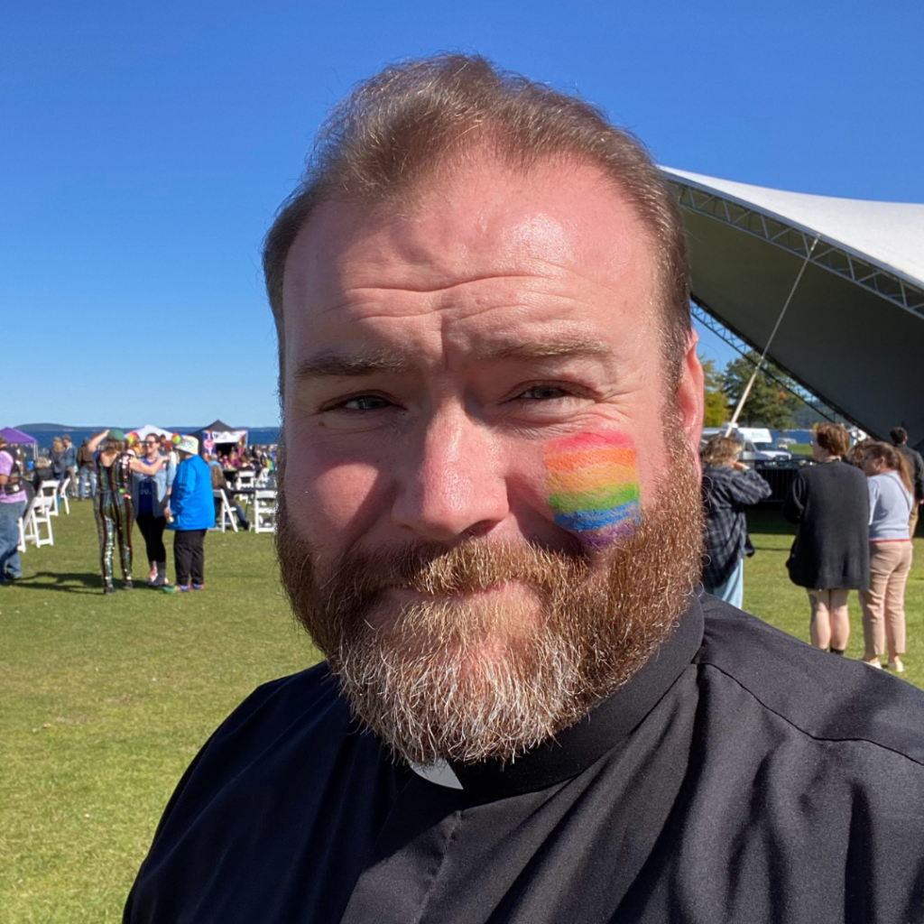 In his clerical collar and shirt and with a rainbow flag painted on his cheek, the bearded Jim Perra smiles to the camera in a selfie at a pride festival.