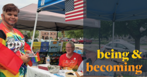 A person in a rainbow cape smiles while handing out items at an outdoor booth with two people seated behind a table. An American flag hangs above, and yellow text reads being & becoming.