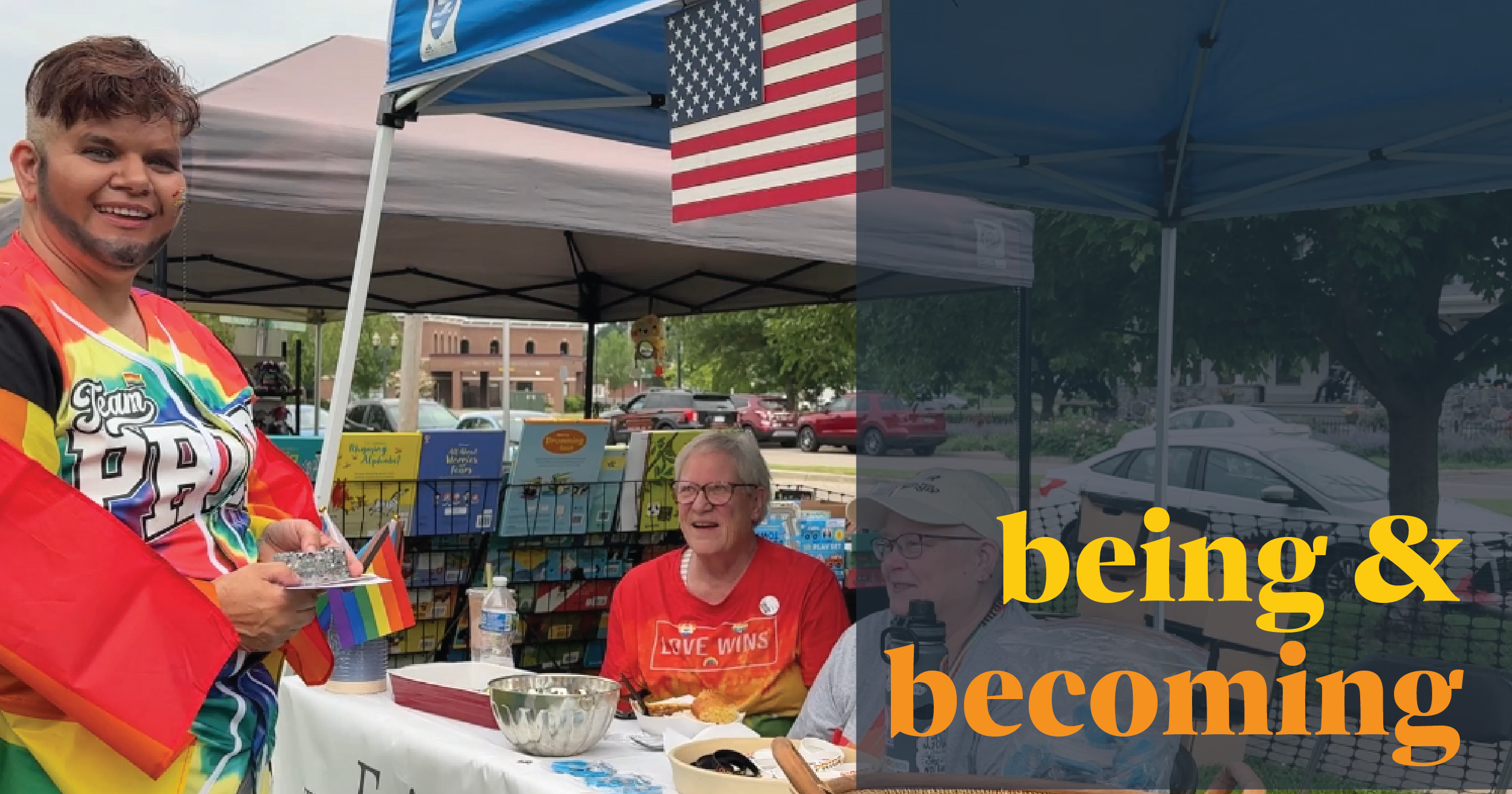 A person in a rainbow cape smiles while handing out items at an outdoor booth with two people seated behind a table. An American flag hangs above, and yellow text reads being & becoming.