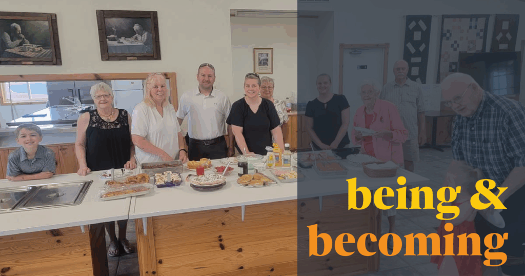 Image Description: A group of adults and two children smile in the parish hall near a generous spread of food. Over the right side of the image is a semi-transparent dark blue color block and text in a bold yellow and rich orange that reads, "being & becoming"
