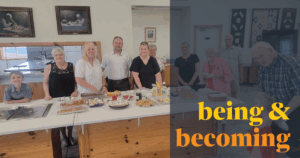 Image Description: A group of adults and two children smile in the parish hall near a generous spread of food. Over the right side of the image is a semi-transparent dark blue color block and text in a bold yellow and rich orange that reads, "being & becoming"