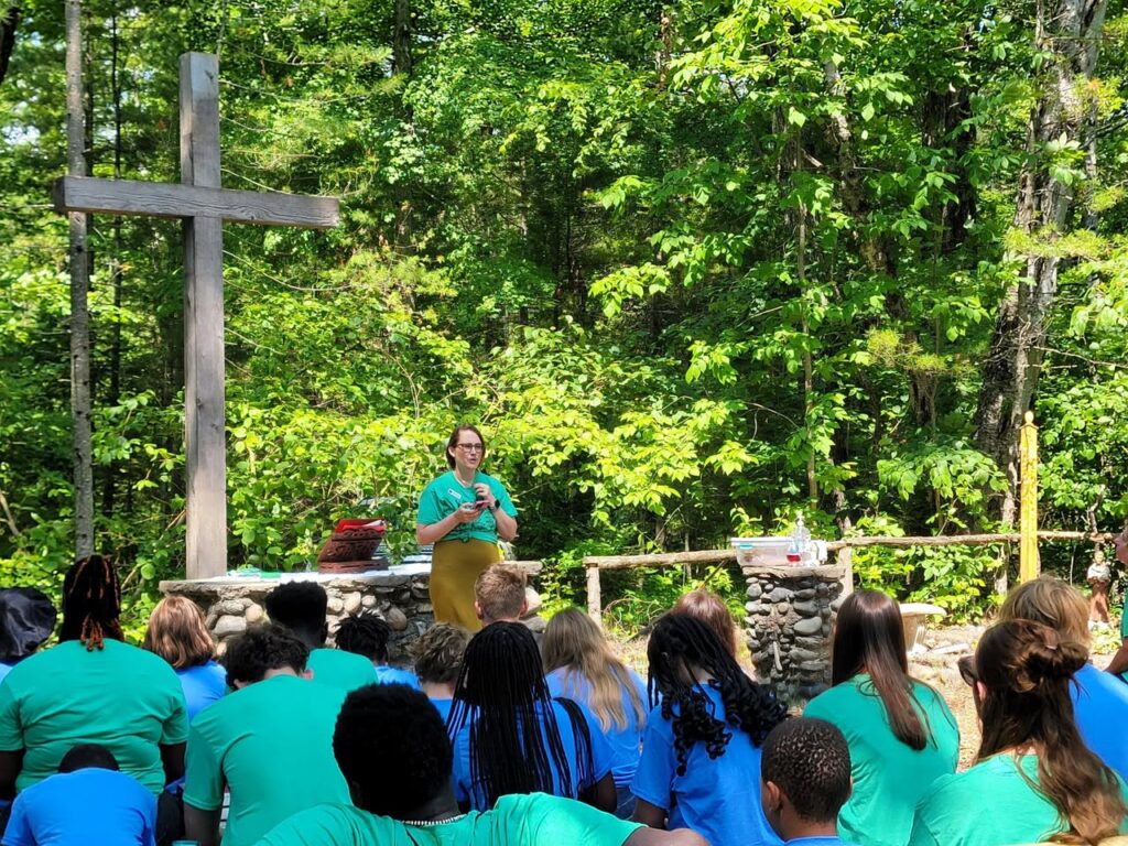 Image Description: McKenize Bade-Knill, Executive Director of Camp Chickagami in Presque Isle, stands in front of the altar of the outdoor chapel in the woods on a sunny day, holding a microphone and addressing the gathered campers, the back of whose heads are in the foreground of the image.