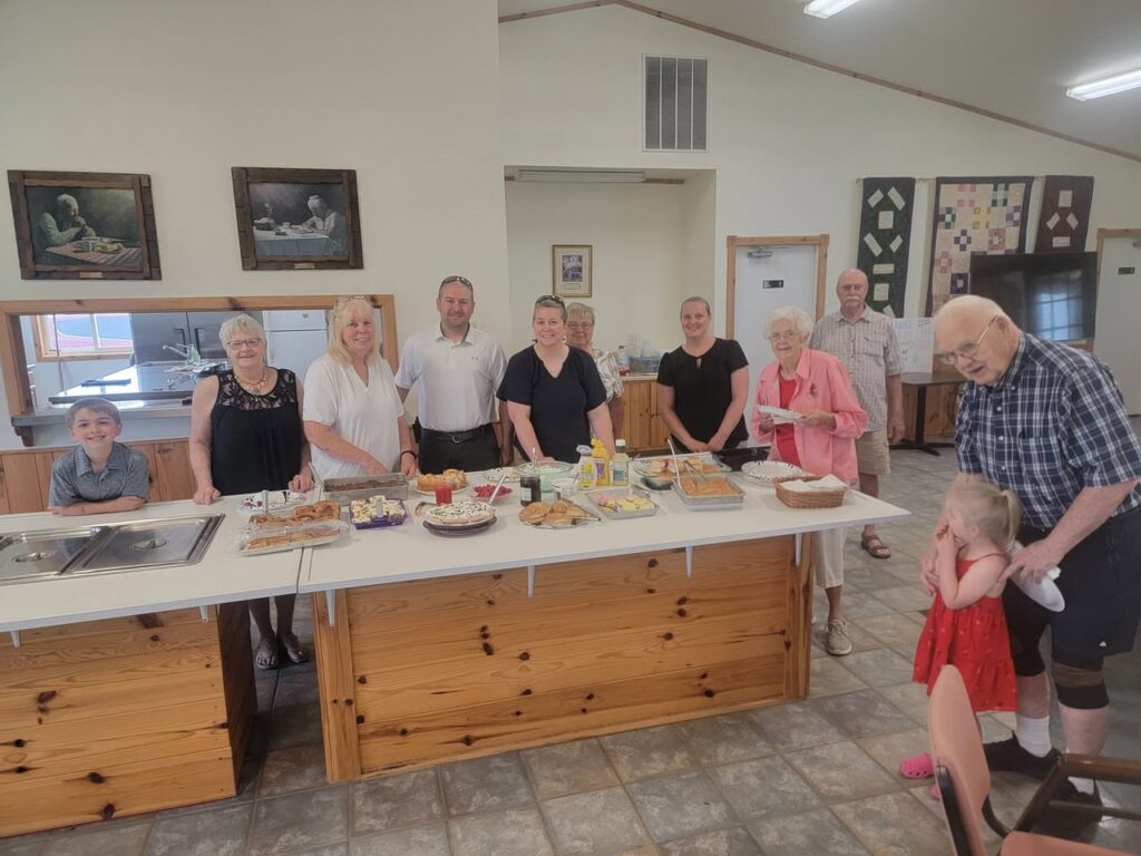 Image Description: A group of adults and two children smile in the parish hall near a generous spread of food. 