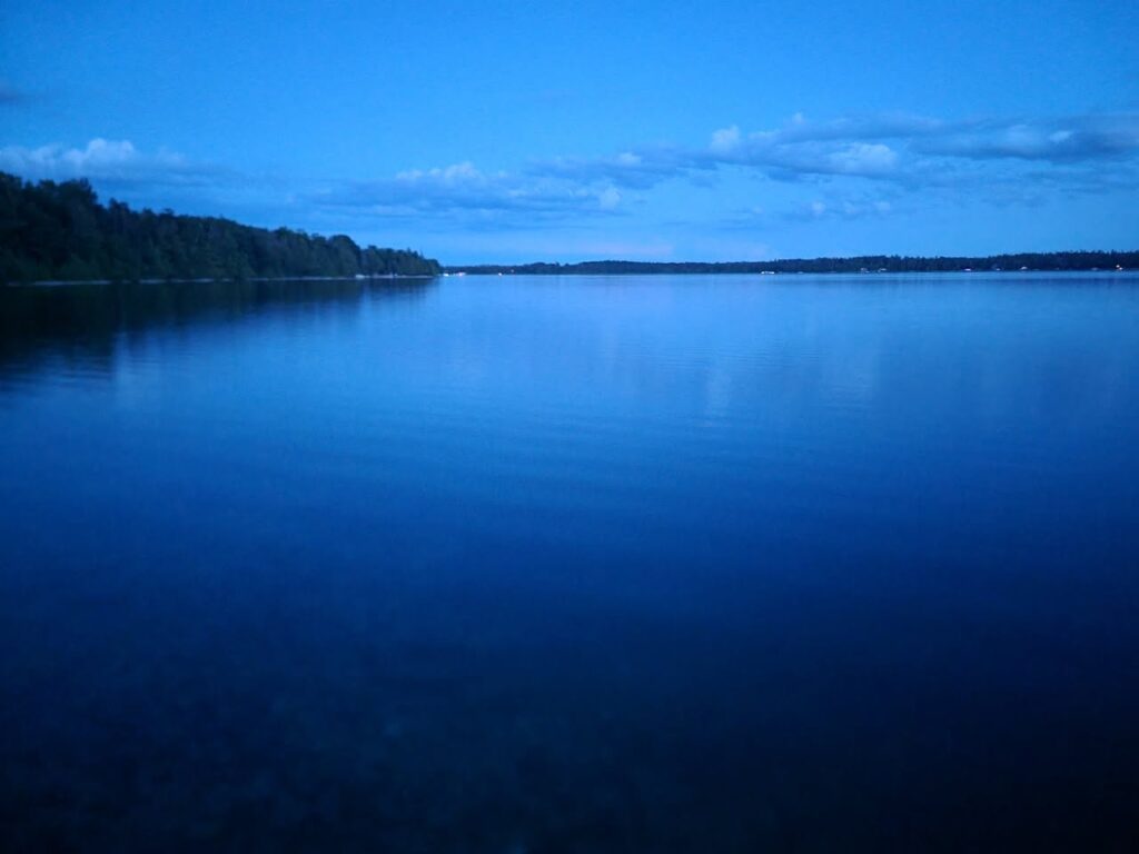 Image Description: Dusk over Lake Esau. The sky is a deep blue with a few low clouds in the distance. The water is as stil as glass and reflects the sky almost like a mirror. On the left side of the image, the shore, comprising pine woods, is visible. 