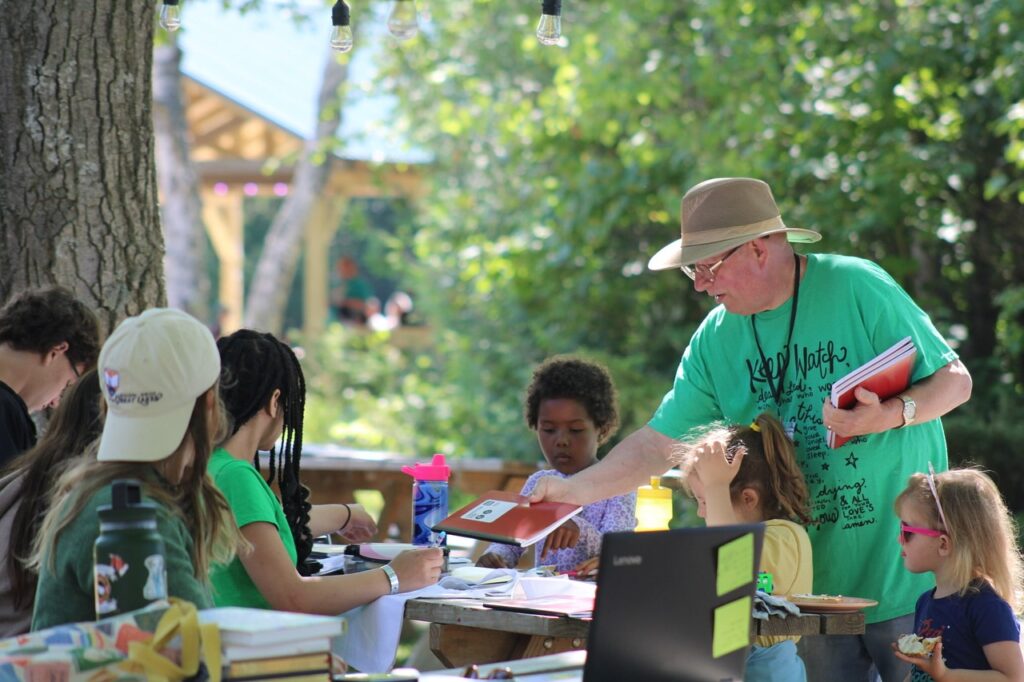 Image Description: Deacon Tom Smith hands out materials to day campers at Camp Chickagami who are seated at picnic tables outdoors on a sunny day. 