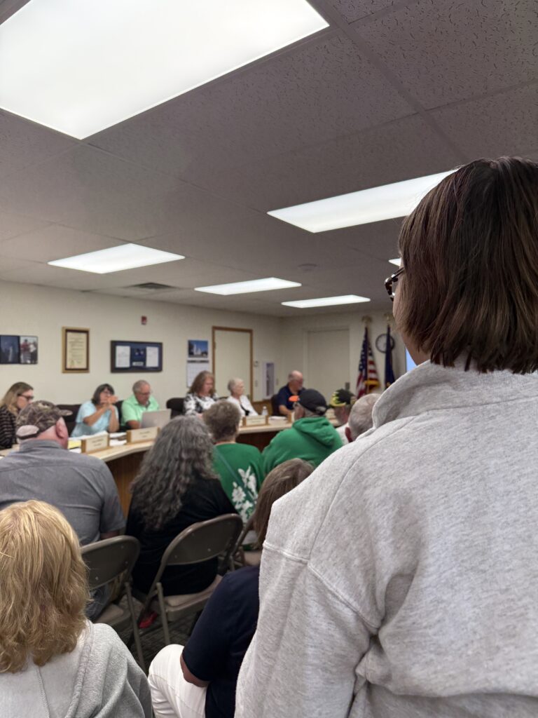 Image Description: The photo is shot from behind McKenzie Bade-Knill who is standing in a Zoning Board meeting, addressing the board members who are seated at a long permanently fixed table at the front of the medium-sized room.