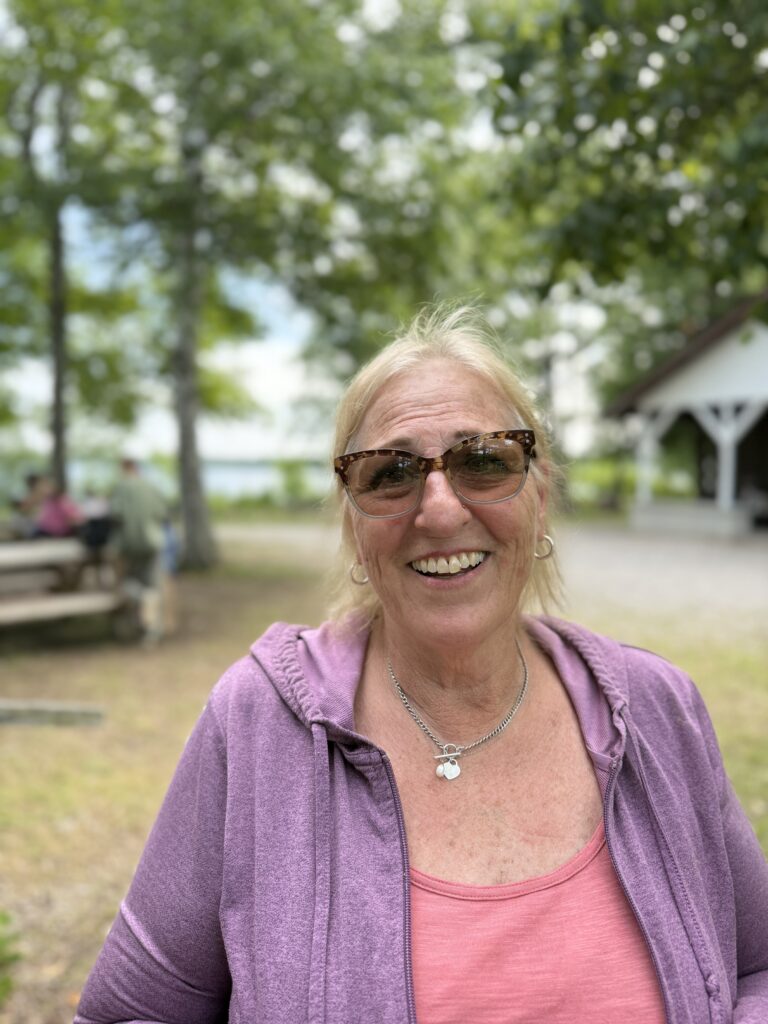 Jeannette Ettin pauses from a busy day at Camp Chickagami to smile for a photo. Camp is just one place where Jeannette's creation care work can be experienced. She can often be found during the busy summer weeks tending to the good news garden or children's learning garden on site. She's dressed in casual clothes and is smiling broadly. The background is in soft focus. It includes a view of Lake Esau, of Fletcher Hall, and campers gathered at a picnic table.
