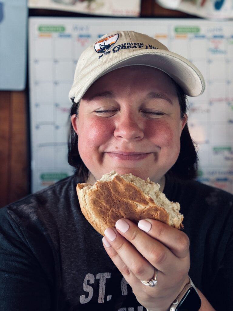 Image Description: A selfie of Rachel Rose-Ravellette. She's wearing a diocesan ball camp and holding a half-gone loaf of fresh bread in front of her face. Her eyes are closed and she's smiling in a way that says, "This is realllllllllly good bread." (It was reallllllllly good. And it was still warm. Deacon Beck made it and he's a fantastic baker.)
