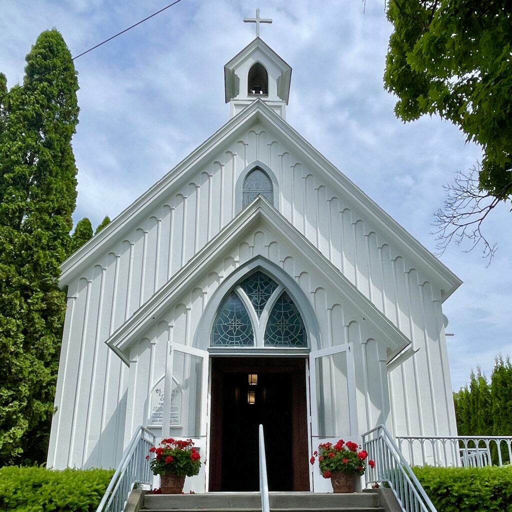 Exterior shot of St. John's, Harbor Springs. It's a white, simple church building with thoughtful details and it has clearly been well loved and carefully maintained. The open, welcoming doors are framed by large planters of stunning red geraniums. The sky is bright blue and the church is surrounded by lush green trees and shrubbery. 