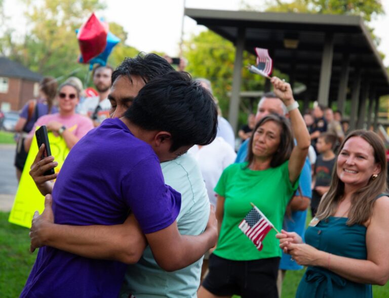 Two people embrace emotionally in a park, surrounded by a smiling crowd. Some people wave small American flags and hold balloons, creating a festive and supportive atmosphere.