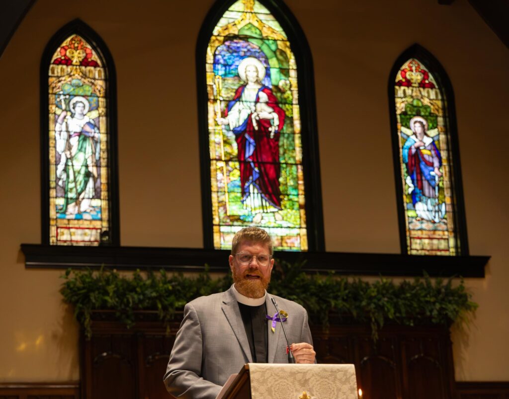 The Rev. Dr. Steven Pankey, in a gray suit, stands at a pulpit at Christ Episcopal Church of Bowling Green Kentucky, with three colorful stained glass windows depicting religious figures behind him.