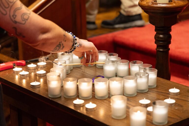 A person with tattoos lights small white candles arranged on a wooden table, using a long wooden stick. More candles and a lighter are visible, with red furniture and other people in the background.