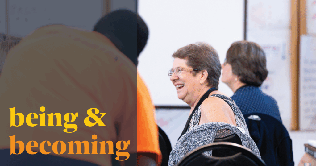 Image Description: In the bright parish hall of St. John's, Midland, during a CCD activity, a participant smiles broadly at someone not pictured. The room is busy and buzzing, as evidenced by the person pictured on the left side of the image in soft focus with his back to the camera who is moving to be part of whatever conversation has left the primary subject so happy. Over the left side of the image is a semi-transparent dark blue color block. In a bold yellow and rich orange, text over the box reads, "being & becoming"