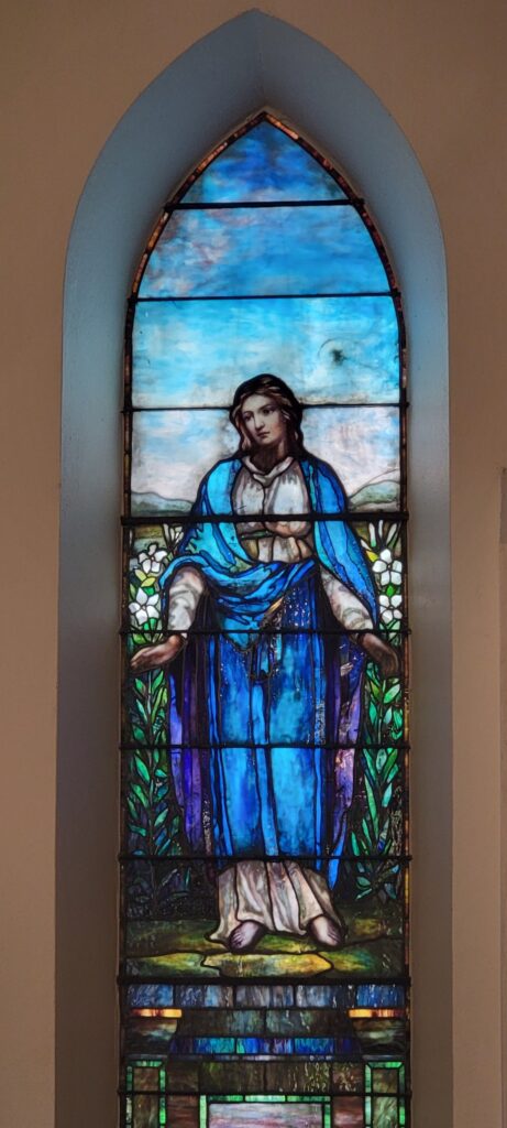 Image Description: A zoomed in shot of a single stained glass window in a church nave. The window is done in a Tiffany Style (it may be a Tiffany window!) and the rich, watercolor-esque colors blend beautifully around the image of a female saint, presumably Blessed Mary, the Mother of our Lord.