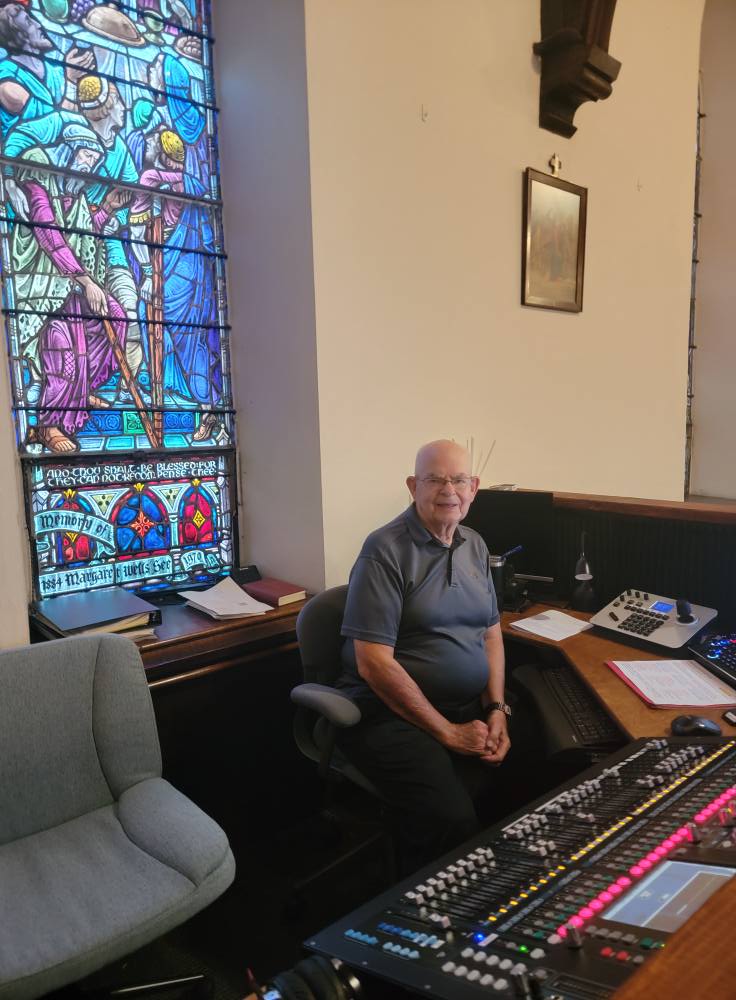 Image Description: An older gentleman sits in an audio/visual booth with a sound board and video mixer in front of him. The booth is in the nave of a church; a colorful stained glass window is behind him.
