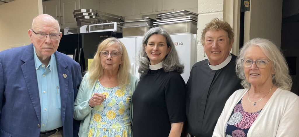 Image Description: A group of five adults stand together in what appears to be the kitchen at Transfiguration Episcopal Church in Indian River. Among the group are Canon Nurya Parish and the Rev. LaRae Rutenbar.