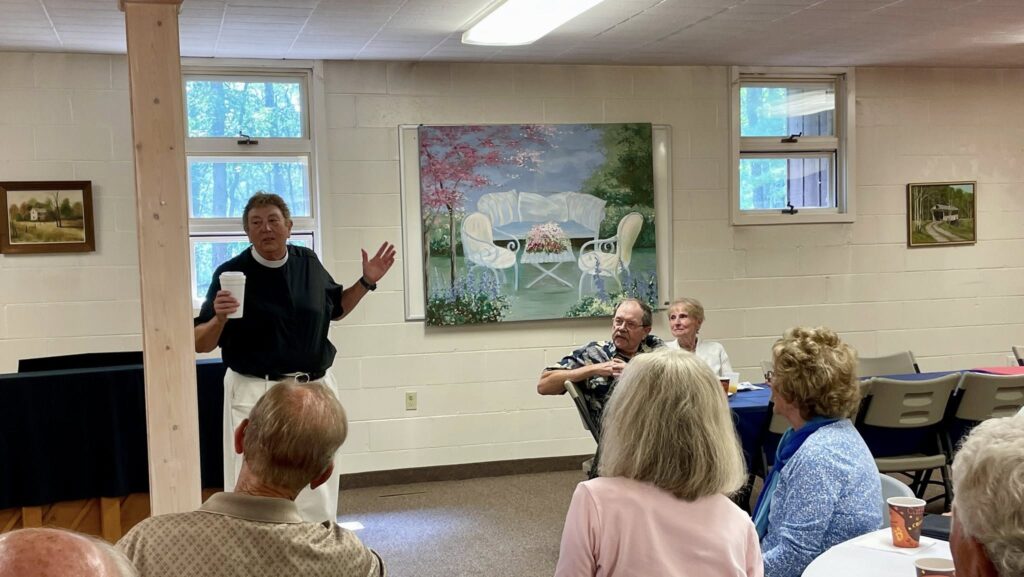 Image Description: In the parish hall at Transfiguration Episcopal Church in Indian River, Rev. LaRae Rutenbar addresses congregants who are facing her while seated at tables during coffee hour.