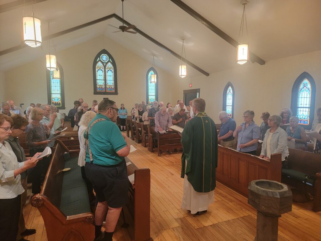 Image Description: A full nave at Epiphany South Haven as shot from the altar. Fr. John Wakefield's back is to the camera, facing the congregation. The nave is simple and bright and you can see the beautifully restored wood floors. John's chasuble is green, the liturgical color of ordinary time.