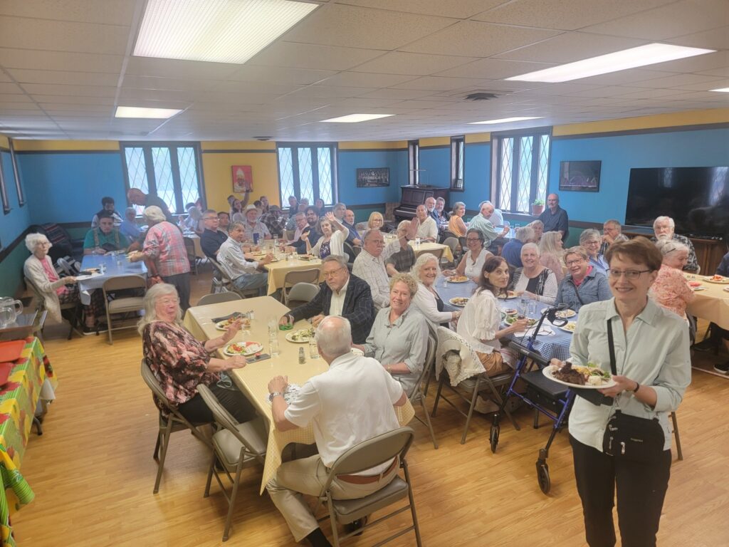 Image Description: A photo of a full-to-bursting parish hall at Epiphany South Haven in which most of the congregants are seated at tables, enjoying brunch together. The room is bright and cheery; it's walls are painted in a vibrant, deep sky blue with deep butter yellow accents.