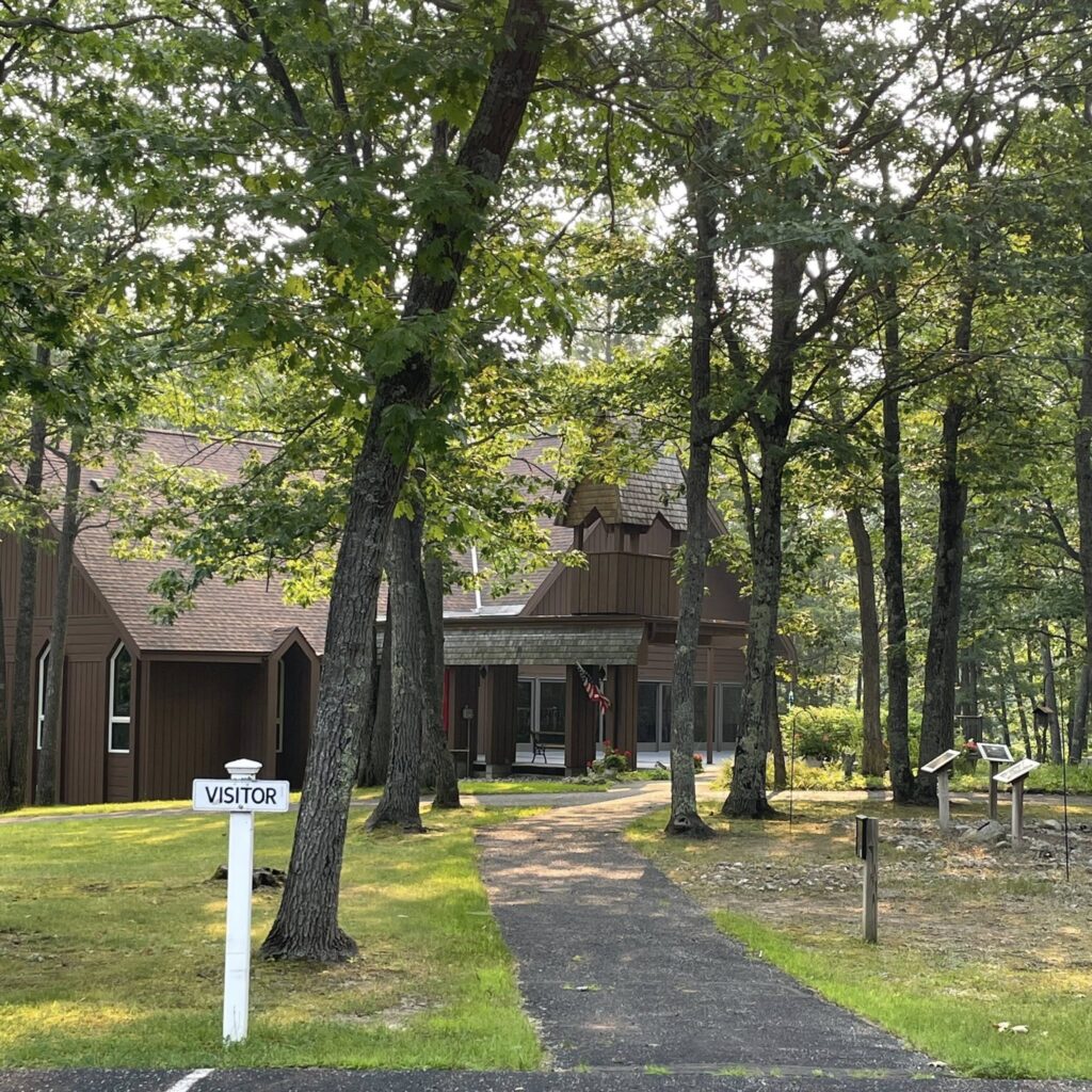 Image Description: An exterior shot of Transfiguration Episcopal Church in Indian River, Michigan. The church is very happily situated in the woods, surrounded by large trees. The building has wood siding, painted a deep, rich brown to blend in with its natural surroundings. You can see a reserved parking sign in the foreground that reads, "VISITOR" and on the right side of the image there is either a garden or a labyrinth marked with stones and placards. The sun is shining through the trees and it is clearly summer - the grass and leaves are verdant.