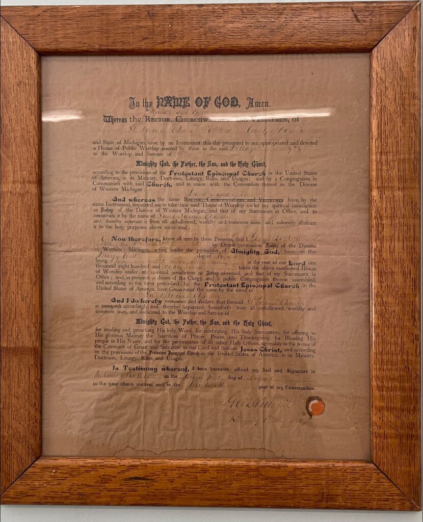IMAGE DESCRIPTION: A close up of a framed, very old, document which appears to be an establishing document for St. James in Pentwater. The date isn't clear, but it's a very old document.