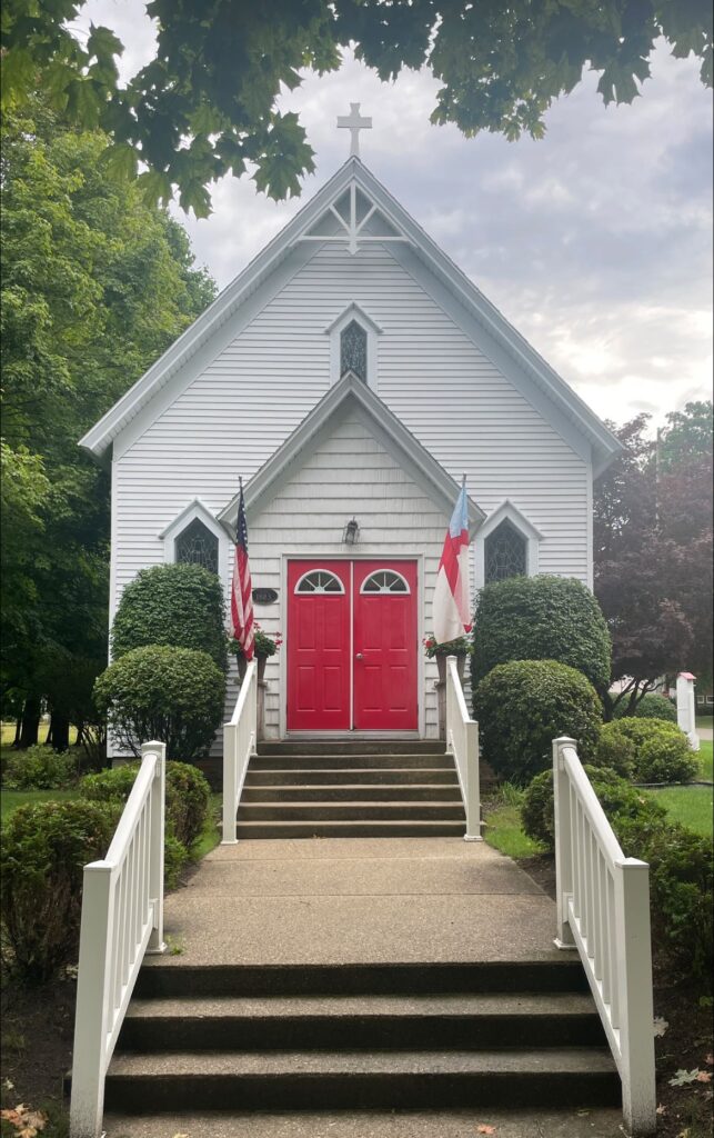 IMAGE DESCRIPTION: Exterior shot of St. James in Pentwater. It's a white clapboard church with double red doors. The sky is blue and the trees are verdant.