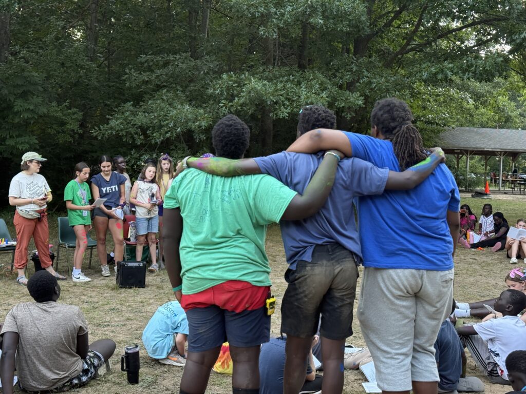 Image Description: Three camp staff members stand with their backs to the camera and their arms around each other as they are led in Compline by a group of campers gathered around a mic and amp. Other campers are seated at the ground, praying along.