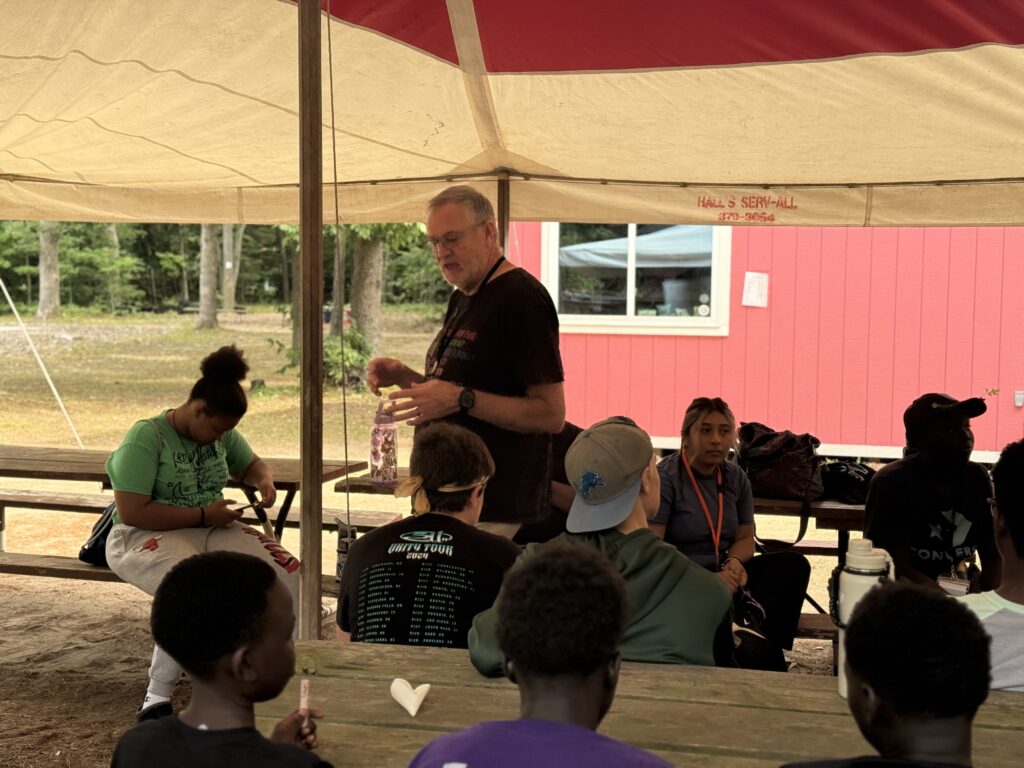 Image Description: Under a large open tent to lessen the impact of the late July heat, Chancellor Bill Fleener teaches campers gathered at picnic tables about collects and the prayers of the people before they took a turn to write their own.