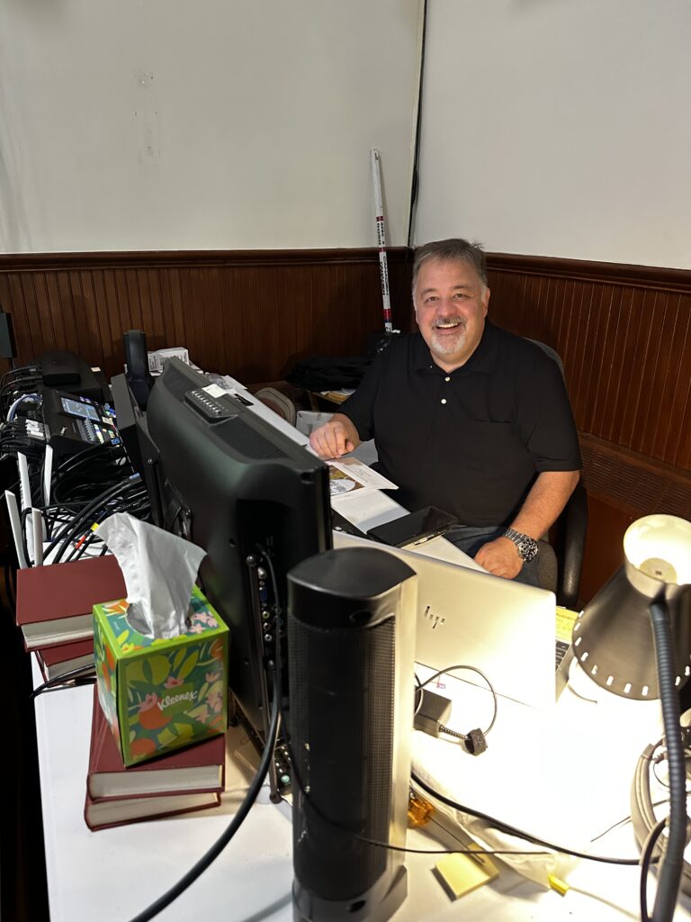 IMAGE DESCRIPTION: A smiling man sits at a tech booth in a church nave.