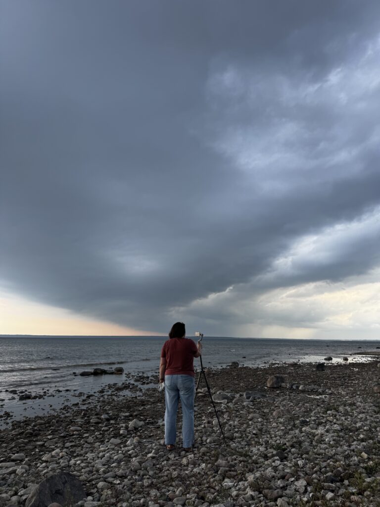 Image Description: A photo taken from behind a person watching a camera on a tripod, taking video of a stunning storm rolling across the straits of Mackinac. The person is standing on a rocky beach and the wind is blowing their hair, but the dark cloud formation is the star of the shot.