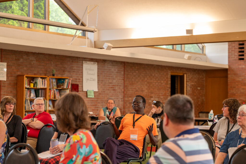 Image Description: In the parish hall of St. John's, Midland, a group of CCD participants are gathered in seats all facing forward. The primary subject of the image is Fr. Zachariah Jok Char who is speaking in response to a prompt to the whole group. Other participants are turned toward him to listen.