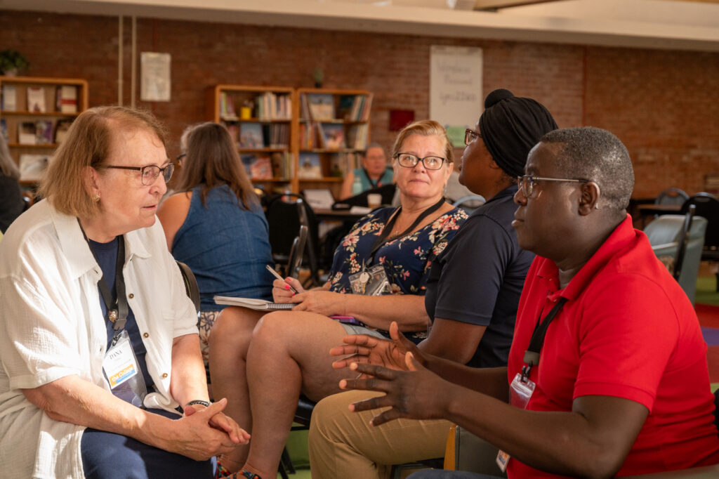 Image Description: In the parish hall of St. John's, Midland CCD participants talk together in dyads and many are pictured in the image. The pair in the foreground are Dana from St. Paul's in Greenville and Fr. Shadrack from Grace in Port Huron. Dana is listening intently to Fr. Shadrack.