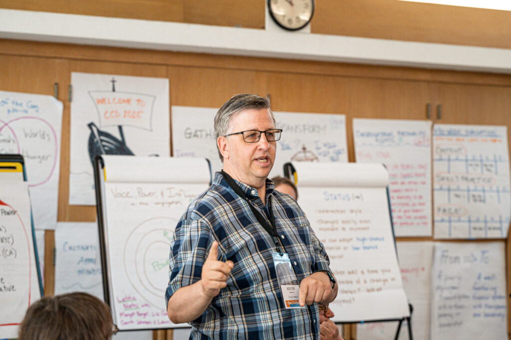 Image Description: Kevin from St. John's in Midland is standing and addressing the room of CCD participants in his home parish's parish hall. Behind him are many easels with large post-in notes with models and other teaching notes in a variety of marker colors. Behind the easels, the wall is covered with even more large post it notes, the signature presentation mode for CCD.