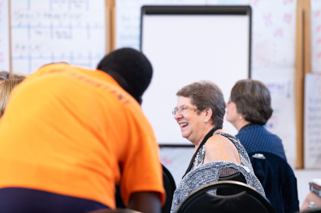Image Description: In the bright parish hall of St. John's, Midland, during a CCD activity, a participant smiles broadly at someone not pictured. The room is busy and buzzing, as evidenced by the person pictured on the left side of the image in soft focus with his back to the camera who is moving to be part of whatever conversation has left the primary subject so happy.