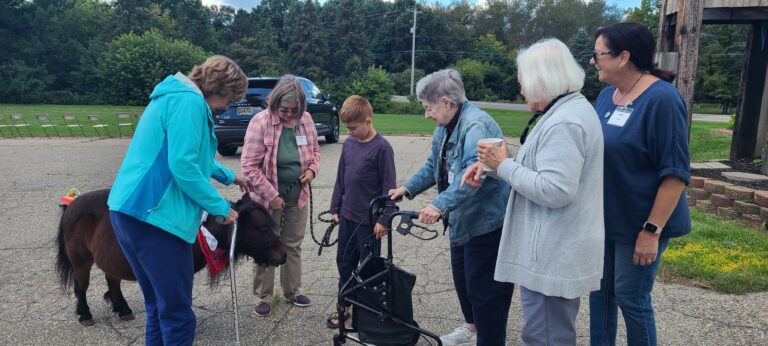 Five women and a young boy stand outdoors, petting a small brown pony. One woman uses a walker, and another holds a cup. They all appear engaged and cheerful on a clear day with trees in the background.