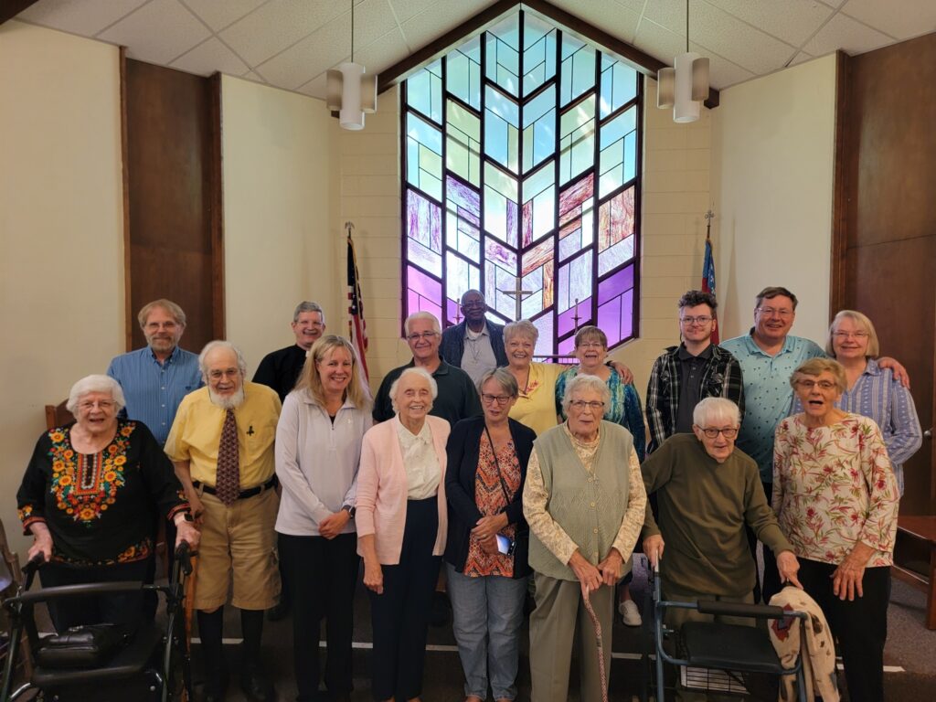 A group of 16 parishioners stand at the front of the nave at St. Barnabas in Portage and smile for the camera. Behind them is a beautiful, geometric stained glass window, the panes of which form a colorful gradient from purple, through mauves, into light greens and blues.