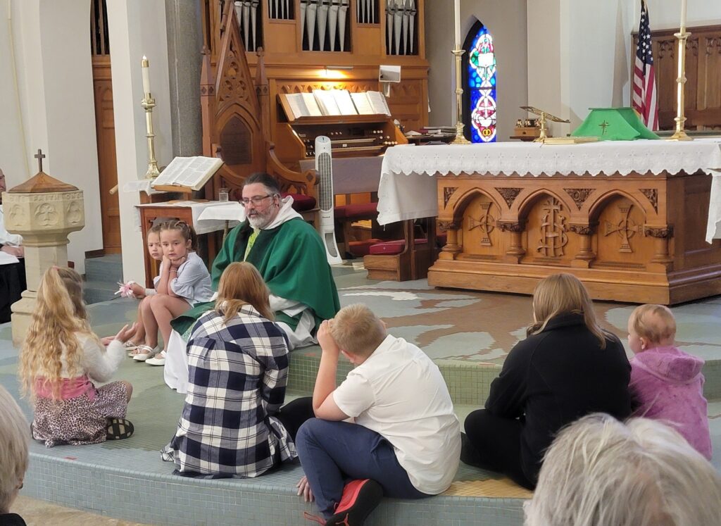 IMAGE DESCRIPTION: Fr. Barrett Lee, vested and wearing a green, ordinary time chasuble, sits on the front edge of the sanctuary in front of the altar with several kiddos on the floor in front of him.
