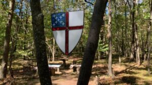The Episcopal Church Shield which is a shield with a red cross and blue quadrant hangs between trees in a sunlit forest. Stone benches and tables are arranged in the background, suggesting an outdoor gathering place.
