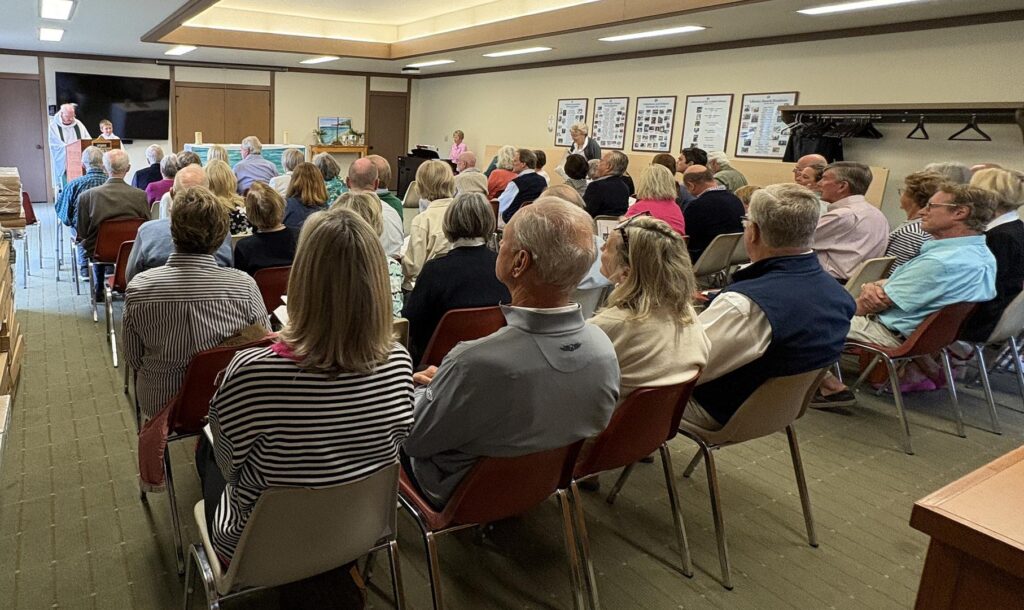 A large group of people are gathered in chairs at the library in Leland where St. Peter's Episcopal Chapel rents space each summer. The room is ful and a priest stands in the front at a lectern.