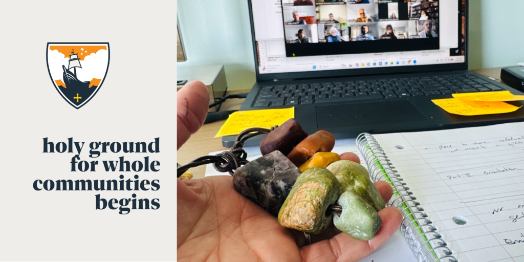 A hand holds several stone pendants over an open notebook with handwritten notes, in front of a laptop showing a video call. Text reads, holy ground for whole communities begins beside a crest logo.