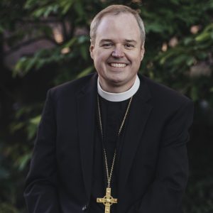 IMAGE DESCRIPTION: A headshot of Presiding Bishop Sean Rowe. He is dressed in clericals and a black sports coat and he wears a large gold pectoral cross on a long chain. He's smiling broadly toward the camera.